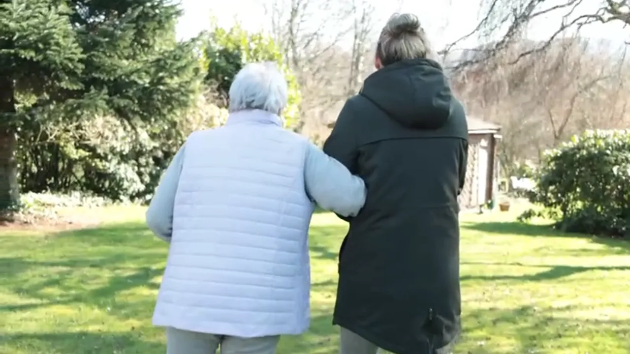 Eine Seniorin geht im Garten Arm in Arm mit einer Begleiterin spazieren